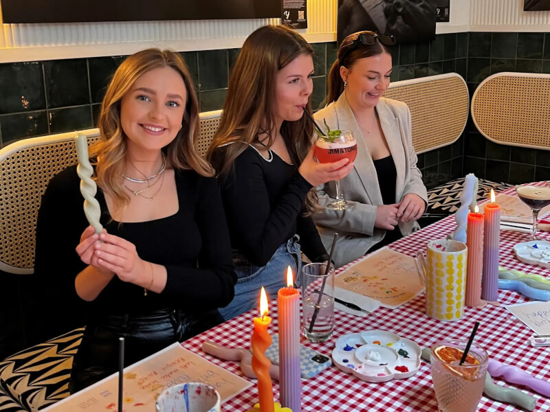 Three women at a Bendy Boy candle making workshop are surrounded by handmade candles and smiling 